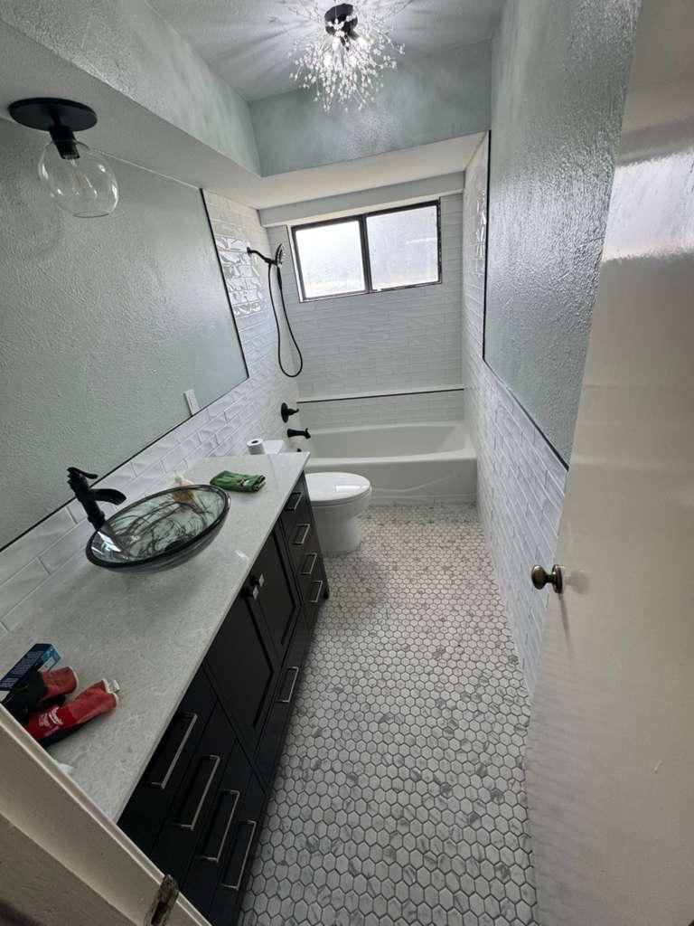 Long white bathroom with a dark vanity, glass vessel sink, small hex floor tile and tub-shower at the back