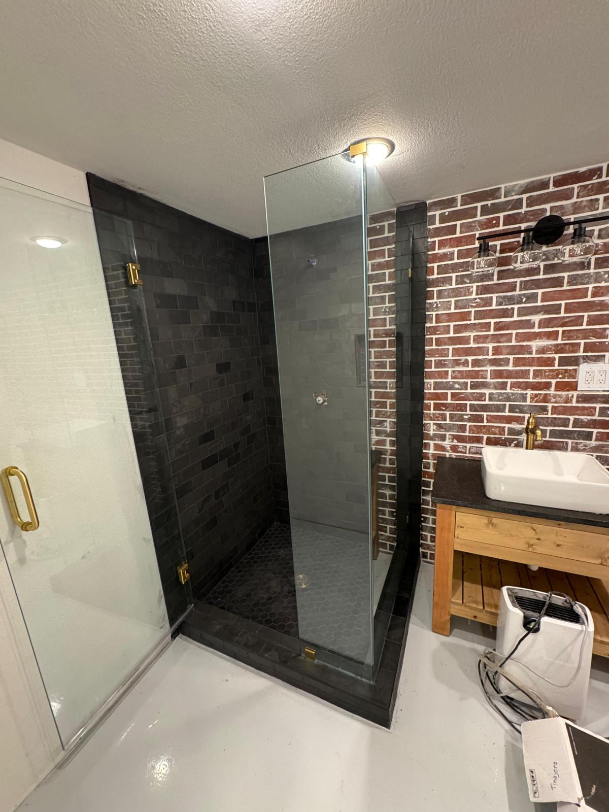 Dark tile shower beside a brick-pattern wall, wood vanity and clear glass partition during finishing work
