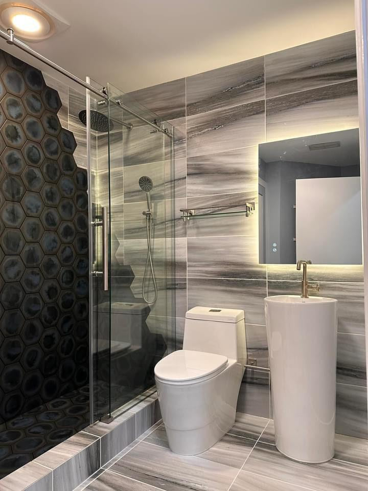 Compact bathroom with gray horizontal tile, a black hex shower wall, clear glass enclosure, toilet and pedestal sink