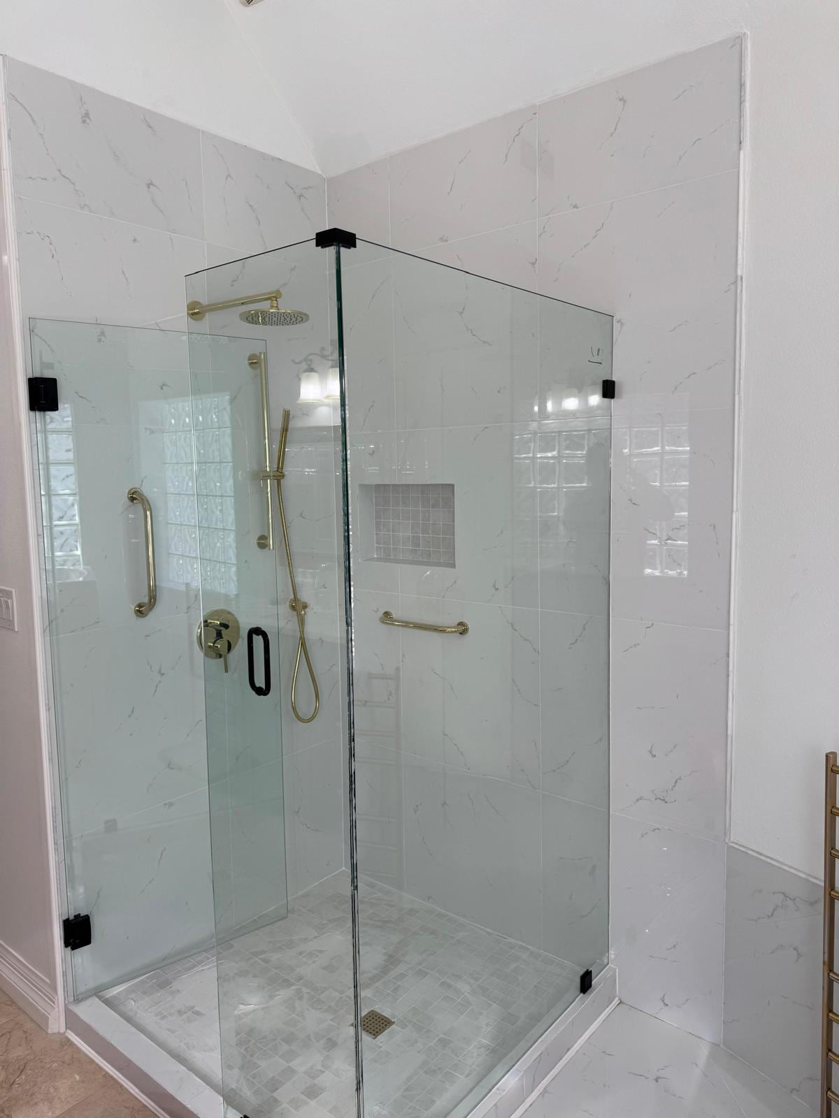 White corner shower with clear glass panels, gold-tone fixtures, grab bars and a recessed niche during finishing work